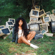 SZA sits on grass, facing the camera. Behind her are various computer monitors and keyboards in a pile.
