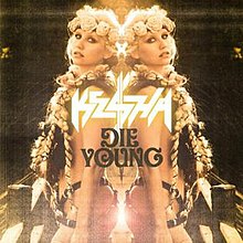 A Caucasian female with blond braids and a floral diadem. She is sparsely clothed in a suit of leather strips. Her back to a mirror, her reflection is symmetrically depicted with the words "Kesha" and "Die Young" appearing in the center.