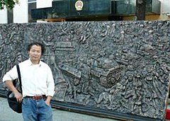 Asian male wearing white shirt and jeans in front of a bronze relief sculpture depicting people, tanks and a statue holding a torch; building with an official Chinese Government emblem is in the background