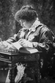 Black and white photograph of a woman seated at an ornate table writing notations in a book.