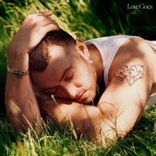 Sam Smith lying down on the ground with their head resting on his left elbow and his right hand resting on his head. A tattoo of two men kissing is visible on his left arm.