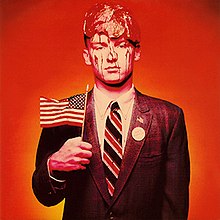 A young man holding an American flag with raw meat dripping on his head and a badge on his chest that reads, "Don't blame me."