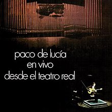 A man plays a flamenco guitar on a chair under a spotlight in a great hall whose back wall is lined with massive organ pipes.