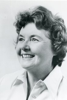 A middle-aged woman with curled hair, photographed in black and white, looking up and to the left, smiling.