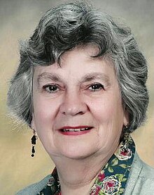 Colour publicity photograph of elderly woman with makeup, permed grey hair and earrings, wearing a chintz-printed blouse and pale jacket.