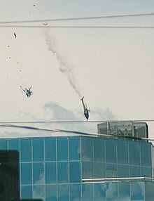 The two helicopters falling mid-air, with the right helicopter, N215TV, emitting smoke, and the left helicopter, N613TV, free-falling upside-down with debris falling above it