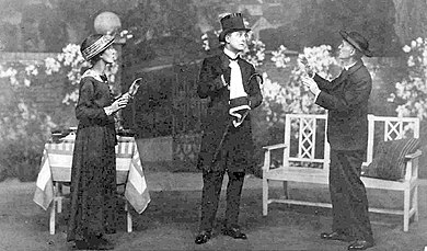 stage scene in a garden setting with a man in full mourning costume centre, older woman to his right and an older man in clerical garb to his left, all wearing hats