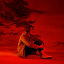 Capaldi sitting with his arms around his legs in a room with red lighting before a red and black background