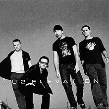 A black-and-white photo of the four members of the band stood next to each other, with Bono crouching while the rest are stood up straight.