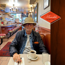 Album cover for Meanwhile (2024) depicting Eric Clapton wearing a bucket hat and sitting in an establishment with a cup of coffee.