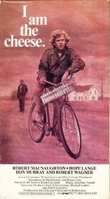 A red-toned scene shows a teenage boy on a bike, one foot down on the path to keep himself still. His left elbow rests on one handlebar while he grips the other. There is a white package in the basket. He looks thoughtful and possibly confused, his brow lowered. Behind him and on the left side of the cover, a man stands in a dark jacket and pants; his face is not visible. On the right, behind both the man and the boy, is a white farmhouse with a black roof. The sparse trees surrounding it look bald. The cover reads "I am the cheese." in white font. The white square below the red cover lists the credits of the film. Most visible are in black at the top of the white square; it reads: "Robert MacNaughton, Hope Lange, Don Murray and Robert Wagner."