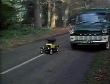 A miniature yellow Ford Model T trundles down a tree-lined country road in sunlight, a full-sized car dwarfing it.