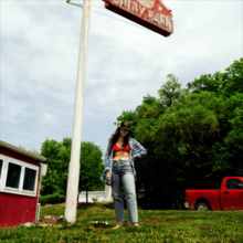 A photo of the artist stood next to a pole holding a sign out of view above her in a grassy field.