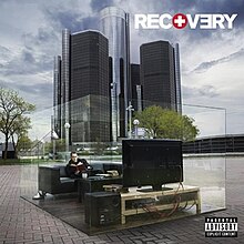 The cover image features skyscrapers in center under cloudy blue sky. Above which, on the ground, transparent glass-type cuboidal body is placed. Inside which, appears a living room with Eminem seated on sofa watching television. On top-left corner, in bold and capitalised format, the title RECOVERY appears.