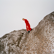 A woman in a red dress (Cate Le Bon herself) walking precariously down the side of a steep boulder.