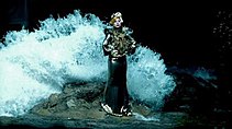A woman stands atop a rock, dressed in a metallic gown and coiffured hair. Behind her, a big wave approaches to engulf her.
