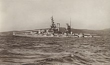 A large light grey battleship tilts back in choppy water as it slowly sinks.