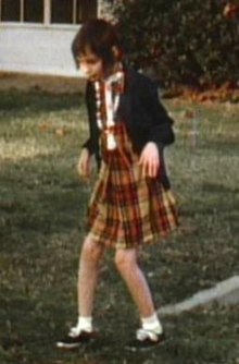 A slightly blurry color picture of Genie, facing slightly right of the camera, walking by herself outside in the hospital recreation yard. She is wearing a red and orange plaid-patterned dress and thin blue sweater, with white socks and black shoes, and looks extremely pale, emaciated, and expressionless. Her limbs are exposed and look extremely thin. Both of her knees are very bent, and her arms are bent forward with both hands hanging down as she holds them out in front of her.