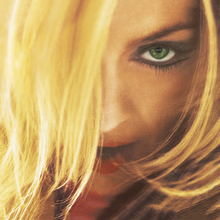Close-up image of Madonna looking to her front and with her opened mouth. She is wearing mascara in her green eyes and her blonde hair hides part of her face.