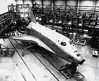 A black and white photo shows a Shuttle-shaped object with a more metallic presentation and missing its rounded nose cone resting in a warehouse surrounded on two sides by long and square metal scaffolding.