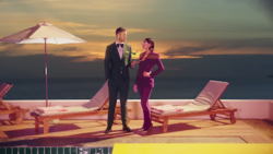 Calvin Harris holding a martini for Dua Lipa on a pool deck. Harris wears a suit and Lipa wears a purple outfit.