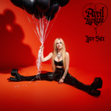 Avril Lavigne sits in the middle of a room with red walls and flooring. She is wearing all black clothing, and holding strings connected to a bundle of black balloons.
