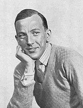 smiling, seated, clean-shaven young white man in tie and jumper, resting chin on hand