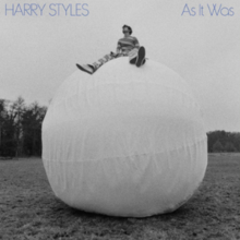 Harry, wearing stripes, sits legs spread atop a large white ball in a field.