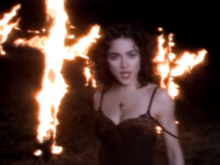 Madonna in front of a pair of burning crosses and is wearing a dark, brown dress, while looking towards the camera