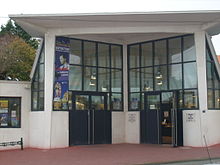 Photographie d'un bâtiment géométrique de béton blanc, une entrée en triangle rentrant avec des hautes verrières et deux portes symétriques de couleur foncée