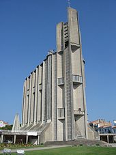 Photographie d'une église moderne en béton brut. Sur trois niveaux, une nef longue et étroite dont les murs côtelés sont ornés de longues bandes verticales faites de carreaux de verre et de balcons aveugles aux striures verticales. Rappelant la forme d'un paquebot se dresse à la « proue » un clocher fait de blocs de béton parallélépipédiques. La base présente des contreforts de béton et un vitrail triangulaire donnant sur un large perron. Un terre-plein couvert de gazon avec des massifs fleuris sépare l'église du parking qui l'entoure.