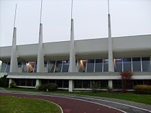 Photographie de la piscine, un bâtiment moderne en béton clair, aux lignes horizontales très marquées, éclairé par de larges baies vitrées. En façade, des sortes de contreforts s'élèvent du sol, allant en s'amenuisant, dépassant la toiture pour se terminer par de hauts mâts métalliques.