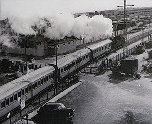 רכבת חוצה את דרך פתח תקווה. המסילה מהקצה השמאלי התחתון של התמונה עד הקצה הימני העליון. הרכבת נוסעת עליה לכיוון ימין התמונה. קרון הרכבת בצד הימני העליון של התמונה, מעלה עשר או קיטור. רכב עומד בימין התמונה למול מחסום סגור, ממתין למעבר הרכבת. תצלום בשחור-לבן.