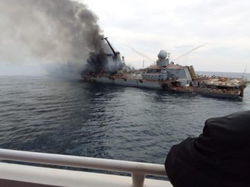 A large warship lists heavily to its port side. Smoke billows from its bridge toward the photographer, who is at the rail of another vessel; the arm of someone else on that vessel is visible in the foreground.]]