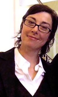Porträtfoto von Sue Perkins. Sie trägt ein schwarzes Jackett, eine weiße Bluse mit aufgeknöpftem Kragen, eine rechteckige, schwarze Brille und ihre schwarzen Haare als bis zum Hals reichende Bobfrisur. Im Hintergrund ist ein braunes Treppengeländer mit weißen Stäben zu sehen.