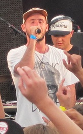 Tom Scott, onstage at a music festival, speaks into a microphone.