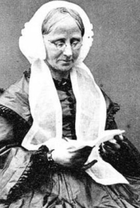 An older white woman seated, wearing a white bonnet with long wide ties; she has grey hair parted center, and is wearing glasses as she looks down into a small book.