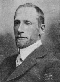 Porträt eines Mannes mittleren Alters mit schmalem Gesicht, kurzen Haaren und Vollbart, er trägt ein weißes Oberhemd, eine helle Krawatte und ein dunkles aber nicht schwarzes Jackett