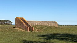 A brick and concrete structure, well-worn, standing on open grass by a concrete track or taxiway