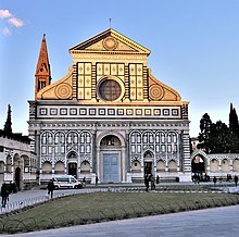 Frontale Farbfotografie einer weißen Kirche mit schwarzen geometrischen Formen. Über dem Rundbogeneingang und beiden kleinen Seitentüren sind Reliefszenen angebracht. In der Mitte der geschwungenen Dachfassade befindet sich ein rundes Fenster und im Dachgiebel erstrahlt eine verzierte Sonne. Links hinter dem Gebäude ist der obere Teil eines braunen Kirchturms zu sehen.