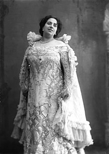 A large woman with dark hair, smiling, standing, wearing a light-colored gown with elaborate beading and a ruffled cape