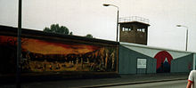 Seitliche Farbfotografie von der Berliner Mauer mit zwei Bildern und einem Wachturm, der hinter dem rechten Bild steht. Das linke Bild zeigt mehrere nackte Menschen und Kinder, die in einem Felsrund mit feuerrotem Himmel im Hintergrund spielen. Das rechte Bild zeigt zwei zusammenlaufende Mauern, die in einem Berg vor weißem Hintergrund mit rotem Kreis enden. An der linken Mauer steht ein Schild „Detour to the japanese sector“.