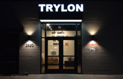 A dark storefront facade with the words "TRYLON" in illuminated white capital letters above the double glass doors. On either side of the doors, lamps cast triangles of light upward and downward to form the shape of an hourglass or bowtie rotated one quarter turn.