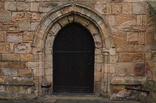 A small window with four leaded panes in a rectangle, another thin pane to the left of thee, in a worn stone-like frame, surrounded by sand coloured stones