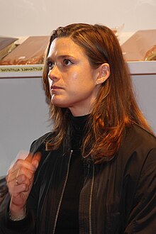 Krista Kosonen seen from profile, wearing a black jacket, looking slightly up with right hand at chest level, in motion