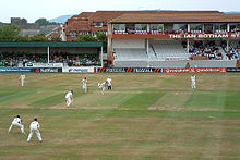 A ground with sportsman playing cricket, with stands in the background