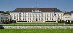 Frontale Farbfotografie von Schloss Bellevue mit einer europäischen und deutschen Flagge im rechten Vordergrund. Ein breiter Rasen mit einer kleinen Mauer als Absperrung führt zu dem zweigeschossigen Gebäude. An das Hauptgebäude sind zwei Seitengebäude angeschlossen. Der Haupteingang hat weiße Säulen und Fensterverzierungen. Im Giebelfeld ist die Aufschrift „Bellevue“.