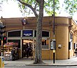 An orange building with an illuminated, blue sign reading "HOLLAND PARK STATION" in white letters and people walking in front all under a white sky