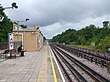 A man with black hair, a black shirt, black pants, and black shoes reading a newspaper and sitting on a brown bench on a grey railway platform