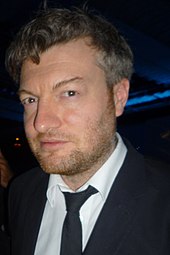 Porträtfoto von Charlie Brooker in einer dunklen Umgebung. Er trägt ein weißes Hemd, ein schwarzes Jackett, eine schwarze Krawatte, einen Dreitagebart sowie seine schwarzen Haare als mittelkurzen Schnitt, wobei einzelne Haare nach oben beziehungsweise unten abstehen.